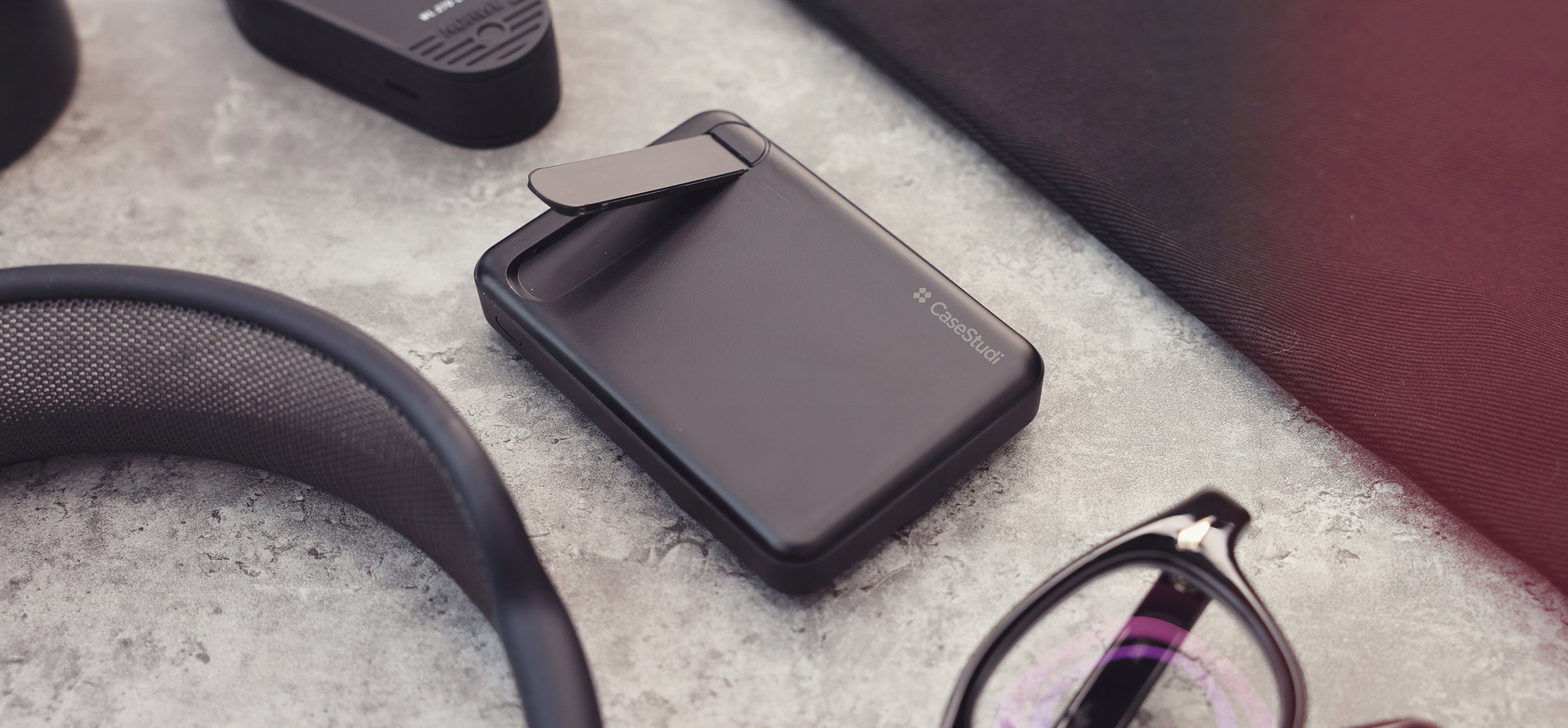 Casestudi FlashMini: 8000mAh power bank with a built-in video stand, lightweight, MagSafe-compatible, and surrounded by daily essentials such as Julius Tart Optical glasses, AirPods Max, and an iPad on a marble table.