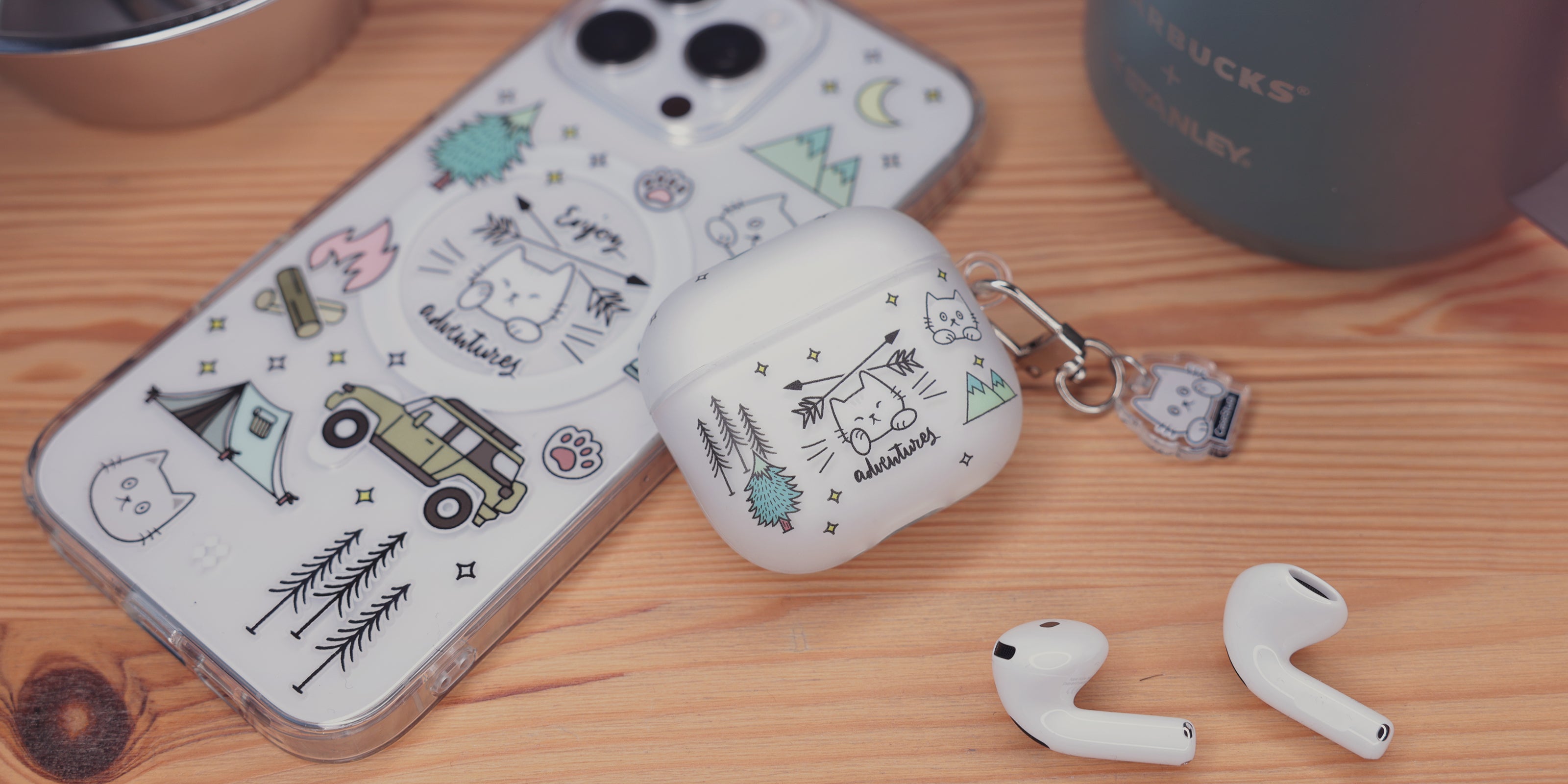 Casestudi AirPods 4 case featuring a camping-themed Cast the cat pattern with illustrations of a jeep, camp tent, campfire, and lantern. The case is placed on a wooden table outdoors, accompanied by a Stanley titanium cup and aluminum cooking tools, capturing the essence of outdoor adventure.