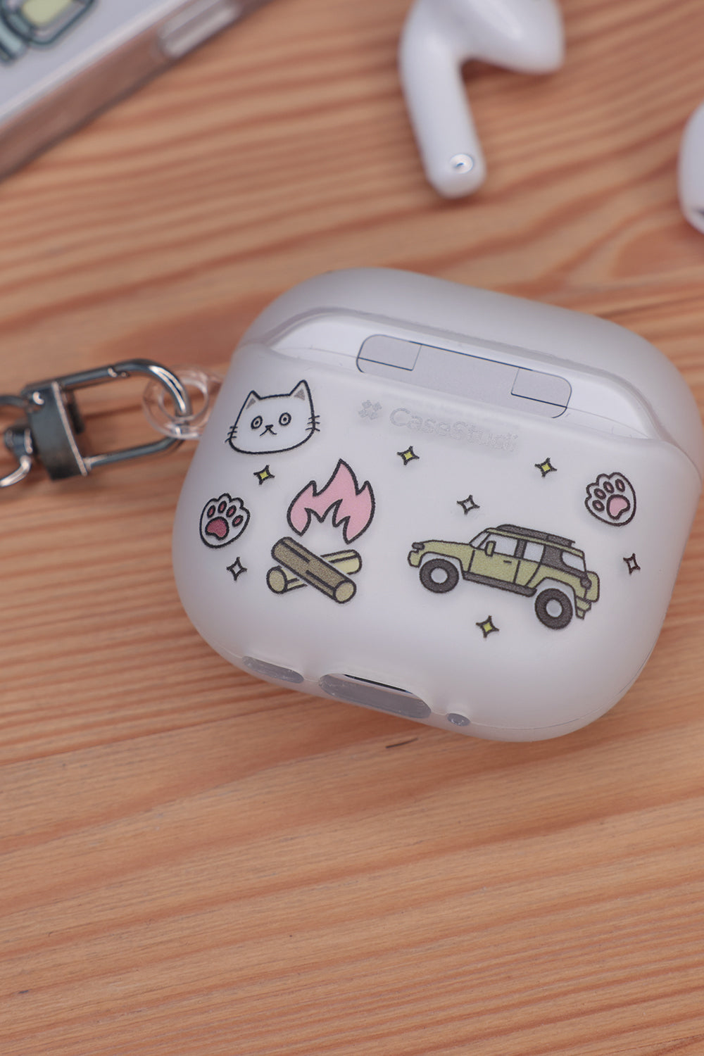 Back view of the Casestudi AirPods 4 case featuring a camping-themed Cast pattern: includes illustrations of a jeep, camp tent and campfire. The case is placed on a wooden table outdoors, complementing the rustic and adventurous vibe.