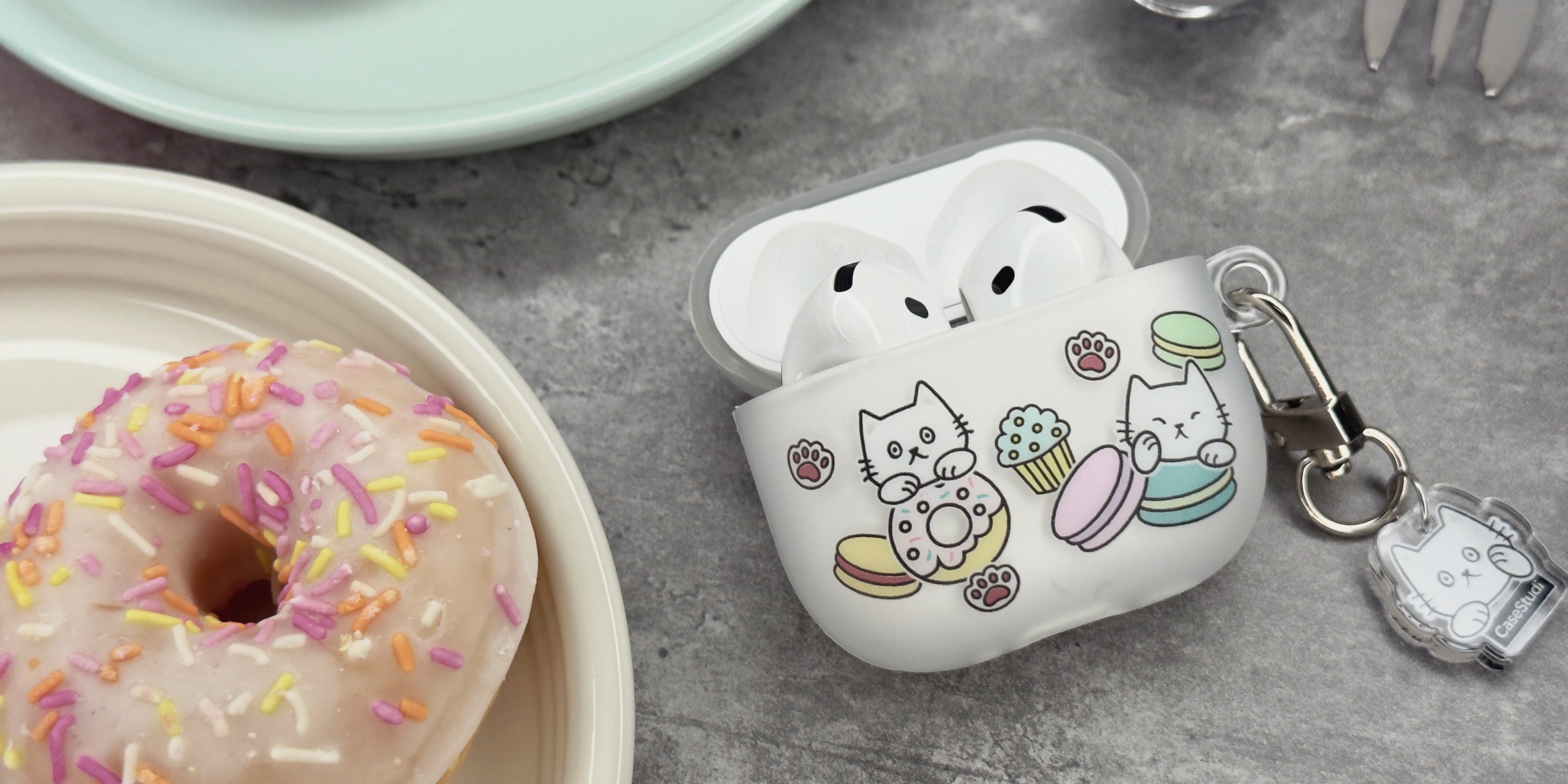 Casestudi AirPods 4 case featuring a Cast pattern: Cast the cat and his friend Woods surrounded by donuts, cupcakes, and macarons. The case is placed on a marble table in a patisserie shop, alongside dishes of colorful macarons and donuts.
