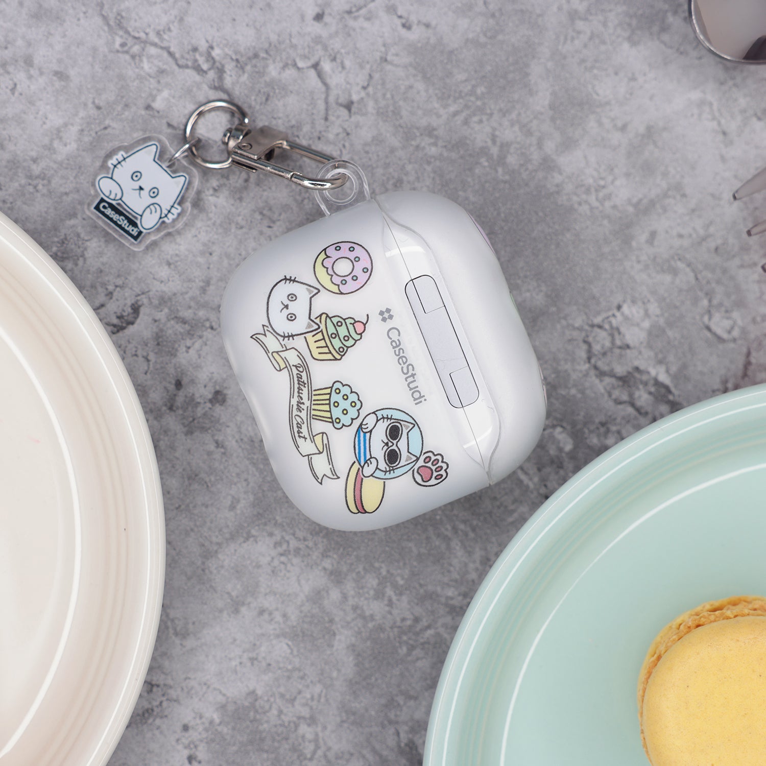 Back view of the Casestudi AirPods 4 case featuring a Cast pattern: Cast the cat and his friend Woods surrounded by donuts, cupcakes, and macarons. The case is placed on a marble table in a patisserie shop, with colorful macarons and donuts in the background.