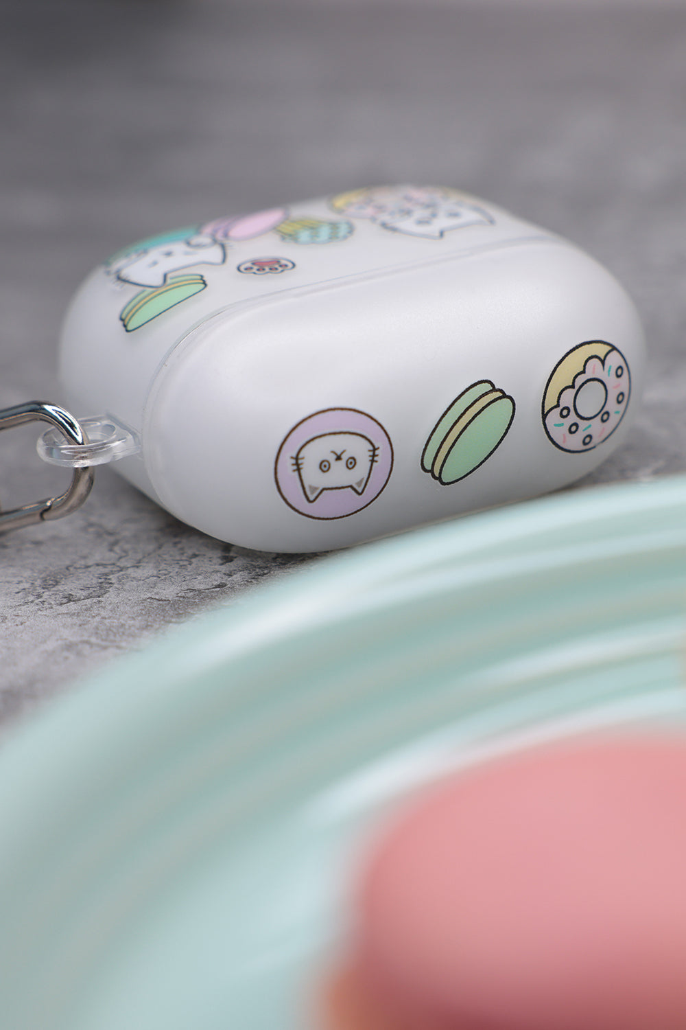 Top view close-up of the Casestudi AirPods 4 case featuring a Cast pattern: Cast the cat and his friend Woods surrounded by donuts, cupcakes, and macarons. The case rests on a marble table in a patisserie shop, alongside vibrant macarons and donuts on dishes.