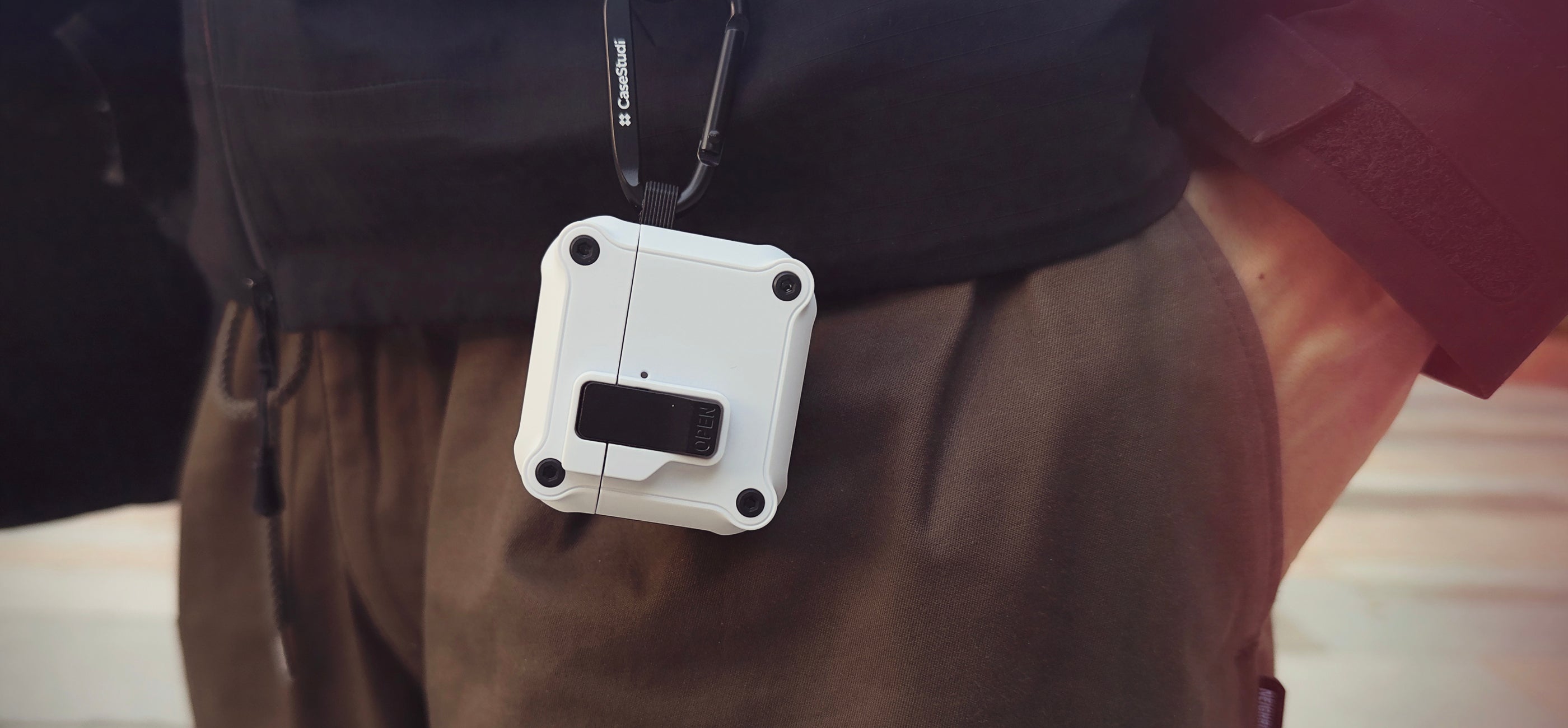 Casestudi white impact case for AirPods 4 with a carabiner, securely clipped to a sleek black Gore-Tex shell jacket. Engineered for ultimate protection with anti-drop durability and a magnetic lock, it’s perfect for keeping your earbuds safe during active days and outdoor adventures.