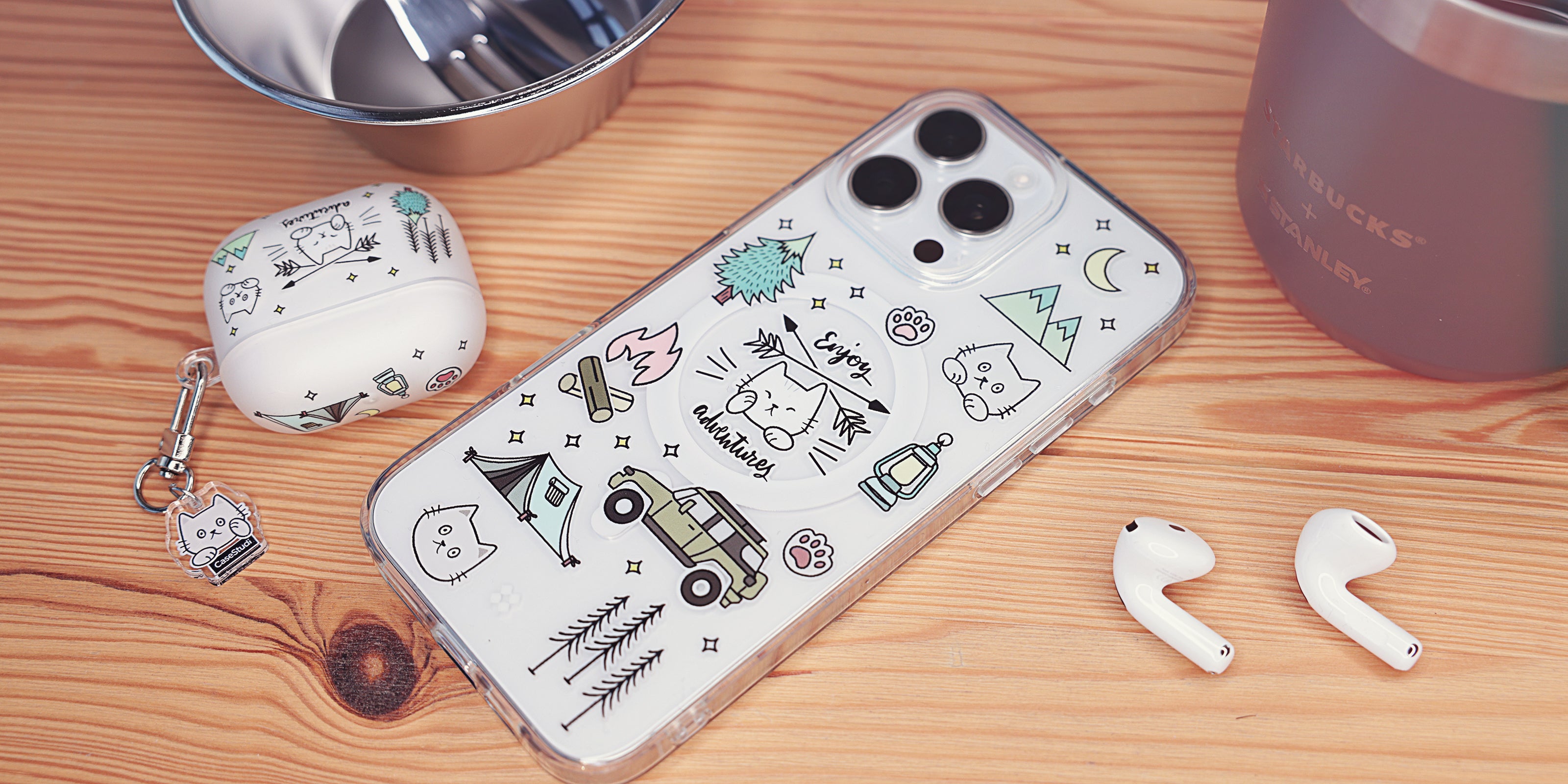 Casestudi iPhone 16 / 16 Pro case featuring Cast the Cat and its friend Woods in an outdoor camping-themed pattern with a forest, jeep, and campfire. The case is placed on a wooden table alongside a Stanley titanium cup, aluminum cooking tools, and a matching Cast-themed AirPods 4 case, creating a cohesive camping vibe.