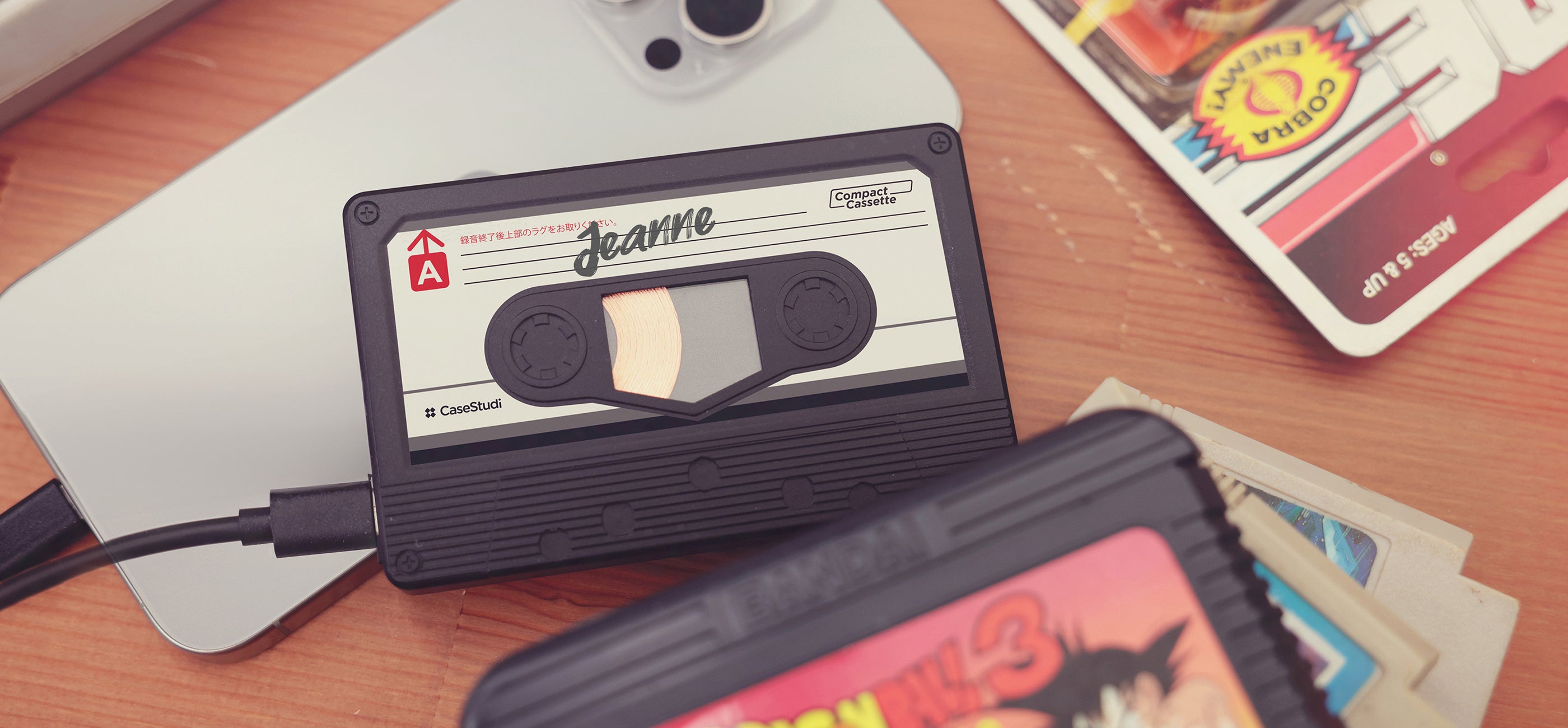 Casestudi Flashtape: A retro cassette-style power bank on an '80s-themed table with vintage items like a Nintendo Dragon Ball game, GI Joe figure, and cassette. Customizable cover and stickers make it a perfect gift for weddings, anniversaries, concerts, or special occasions. Deliver a message to your loved ones or express yourself with your own design, combining nostalgia with advanced charging for smartphones, Switch, and tablets.