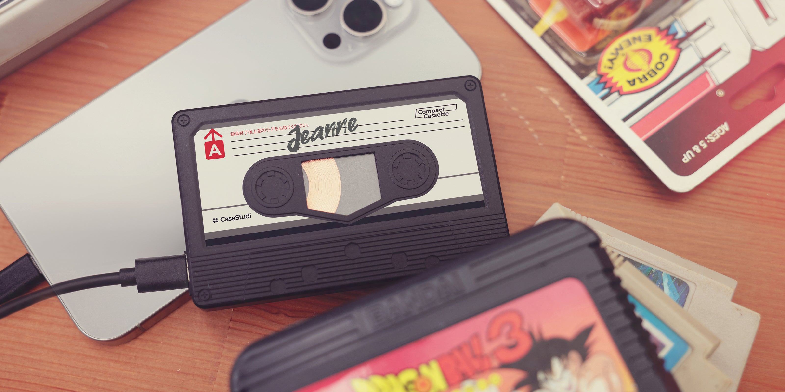 Casestudi Flashtape: A retro cassette-style power bank on an '80s-themed table with vintage items like a Nintendo Dragon Ball game, GI Joe figure, and cassette. Customizable cover and stickers make it a perfect gift for weddings, anniversaries, concerts, or special occasions. Deliver a message to your loved ones or express yourself with your own design, combining nostalgia with advanced charging for smartphones, Switch, and tablets.
