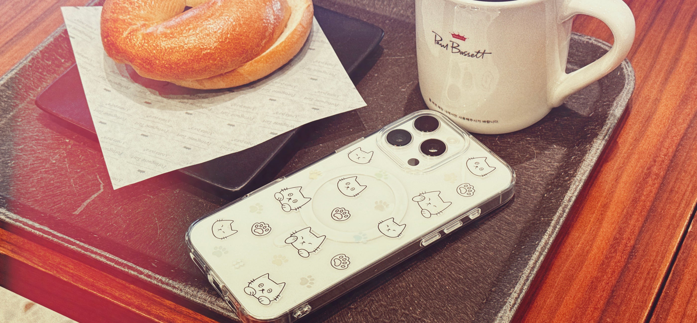 Casestudi iPhone 16 Pro Max Case with Cast character and cat footprints, showcased in a cozy café setting with a bagel and coffee, perfect for cat lovers who appreciate a relaxed and stylish vibe.