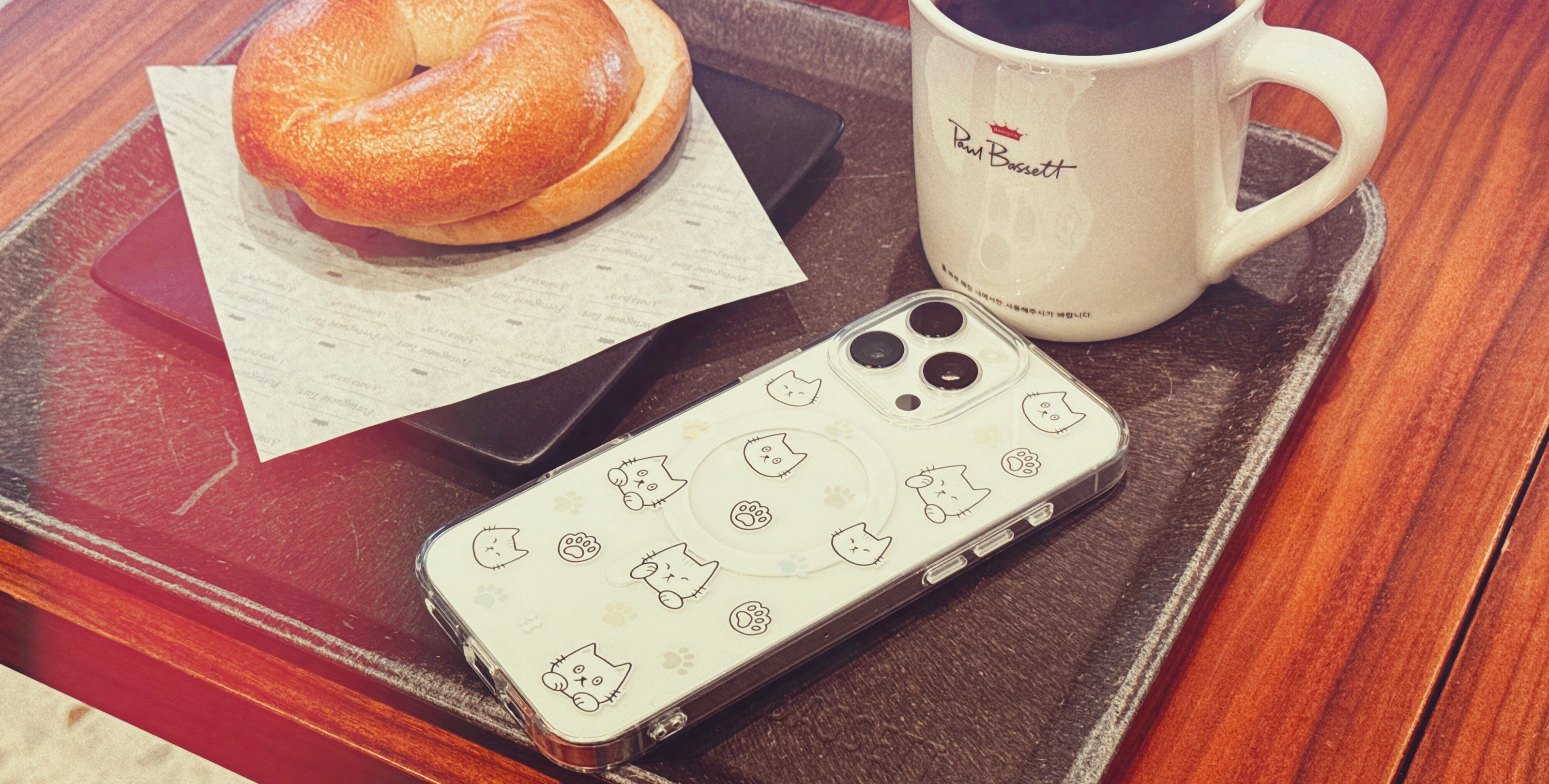 Casestudi iPhone 16 Pro Max Case with Cast character and cat footprints, showcased in a cozy café setting with a bagel and coffee, perfect for cat lovers who appreciate a relaxed and stylish vibe.