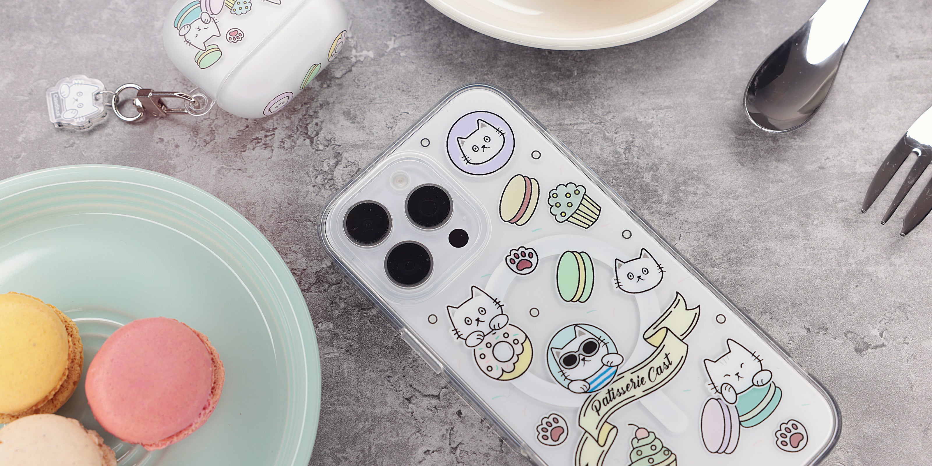 專為 iPhone 16 Pro / 16 設計的 Casestudi Cast the Cat 保護殼，呈現 Cast 貓咪圖案，周圍環繞著甜甜圈、杯子蛋糕與馬卡龍，搭配柔和的馬卡龍色調，展現可愛與溫馨的設計風格。保護殼放置於大理石桌面上，旁邊擺滿甜點盤，完美襯托出甜美又精緻的氛圍。