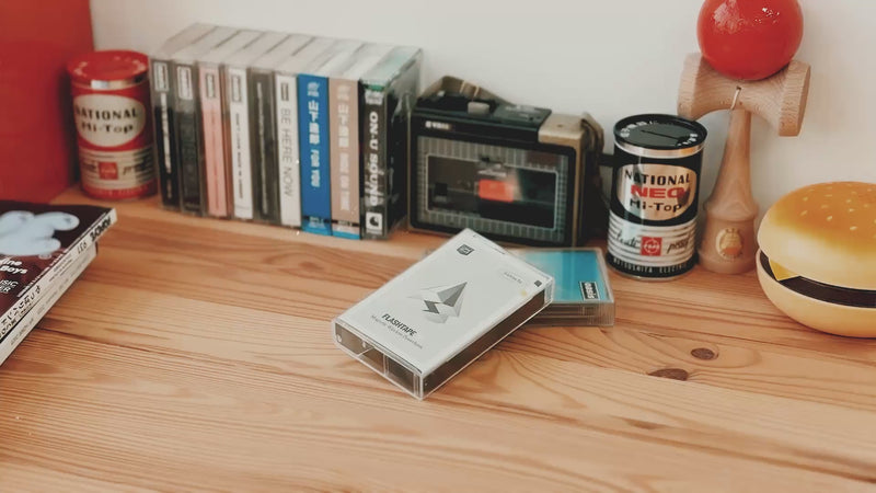 Casestudi Flashtape EP unboxing video set in a retro-inspired scene. The unboxing takes place on a wooden table with nostalgic items like Oasis cassettes, an 80s hamburger-shaped retro phone, and an old-school cassette player. The Flashtape EP comes in a real cassette box, revealing a cassette-shaped 5000mAh power bank. It includes stickers for customization, letting you write messages and relive the charm of the good old days.